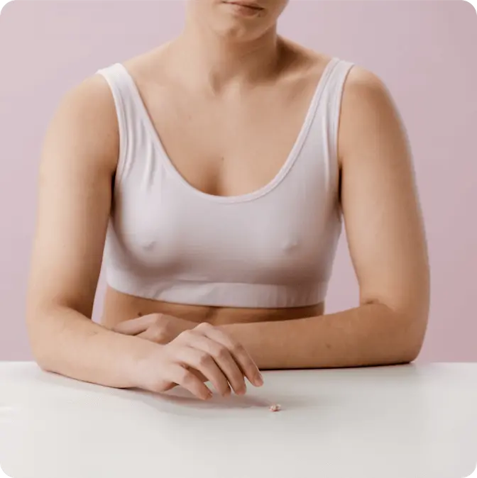 Человек в белом спортивном бюстгальтере сидит за белым столом на нежно-розовом фоне, глядя на конфету. Атмосфера спокойная и созерцательная.